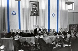 David Ben-Gurion (First Prime Minister of Israel) publicly pronouncing the Declaration of the State of Israel, May 14 1948, Tel Aviv, Israel, beneath a large portrait of Theodor Herzl, founder of modern political Zionism, in the old Tel Aviv Museum of Art building on Rothshild St. The exhibit hall and the scroll, which was not yet finished, were prepared by Otte Wallish.  Photo by Rudi Weissenstein - Israel Ministry of Foreign Affairs, Public Domain, https://commons.wikimedia.org/w/index.php?curid=39649093